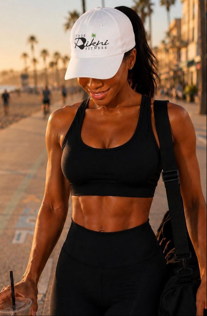 A woman walking on a beach boardwalk at sunset wearing a white baseball cap with a black logo, a black sports bra, and black leggings, with palm trees and ocean in the background.